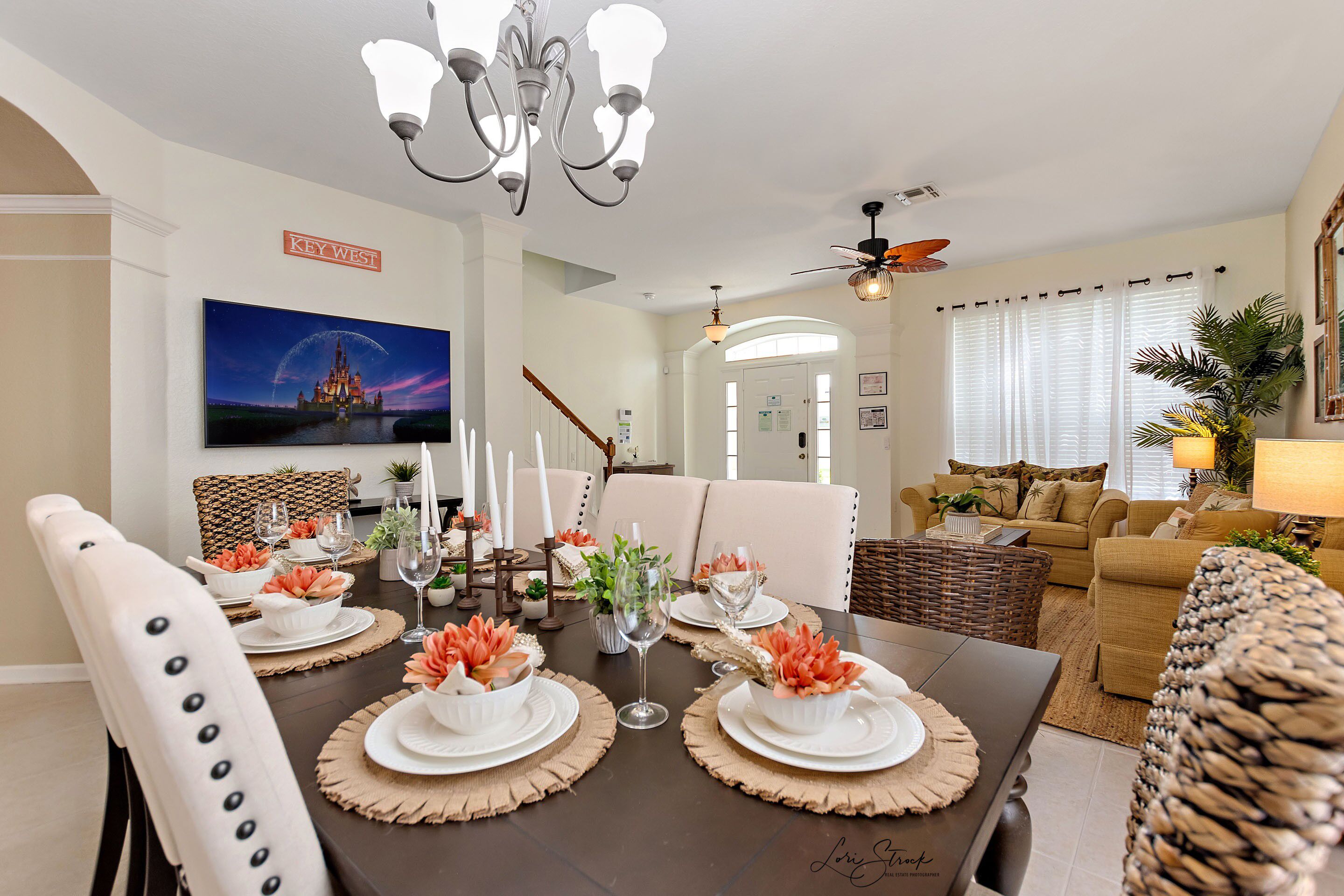 A dining area with a dark wooden table set for four, white plates, and orange flowers. There is a chandelier above the table and a ceiling fan in the background.
