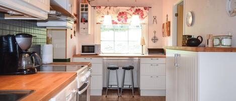 A compact kitchen with a wooden countertop, a microwave, a kettle, and a coffee maker. There are two stools and a window with a floral curtain.