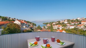 Un balcon avec une table dressée pour deux personnes, donnant sur un village côtier aux maisons aux toits rouges et sous un ciel d’un bleu limpide.