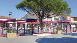 A row of outdoor dining areas with red awnings, a large tree in the center, and buildings in the background.