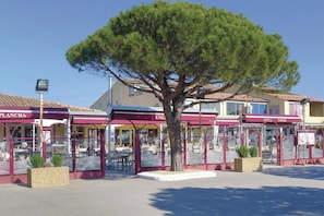 A row of outdoor dining areas with red awnings, a large tree in the center, and buildings in the background. - Homerez - Apartment Port-de-Bouc with Pool (Port-de-Bouc)