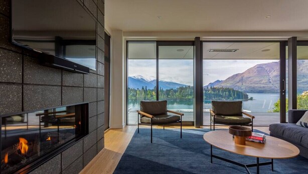 A modern living room with a fireplace, two armchairs, a coffee table, and a large glass door leading to a scenic outdoor view.