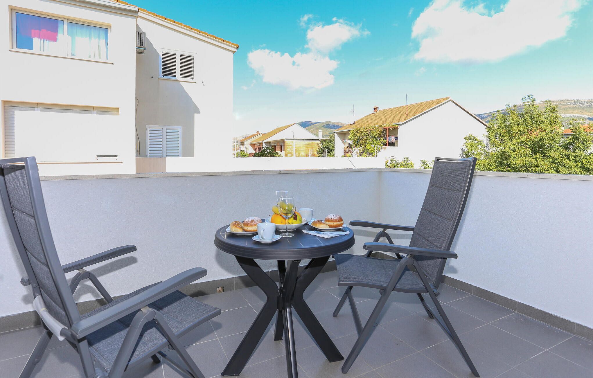 Uma pequena mesa redonda com duas cadeiras numa varanda com parede branca e vista para casas ao fundo.
