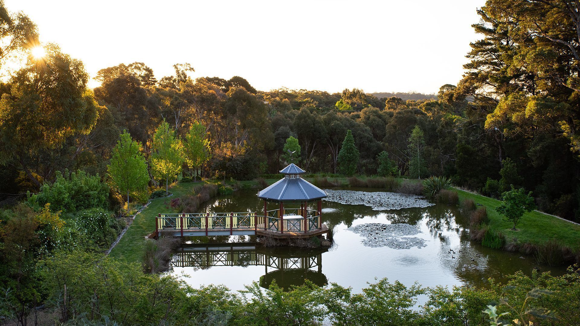 A gazebo with a thatched roof and a wooden deck over a pond, surrounded by lush greenery and trees.