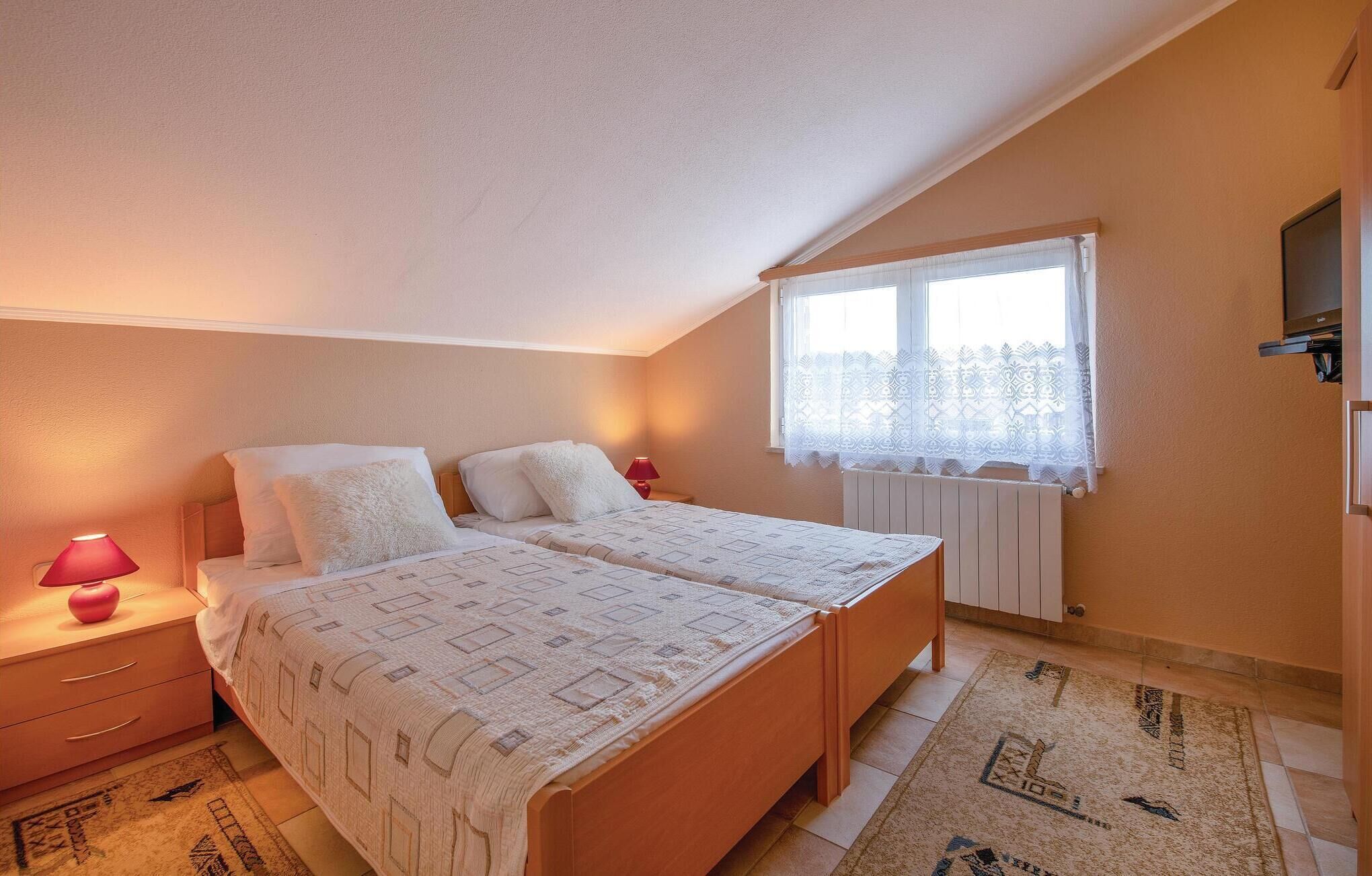 A bedroom with two beds, a window with lace curtains, a radiator, and a TV mounted on the wall.