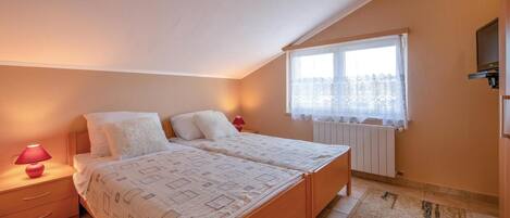 A bedroom with two beds, a window with lace curtains, a radiator, and a TV mounted on the wall.