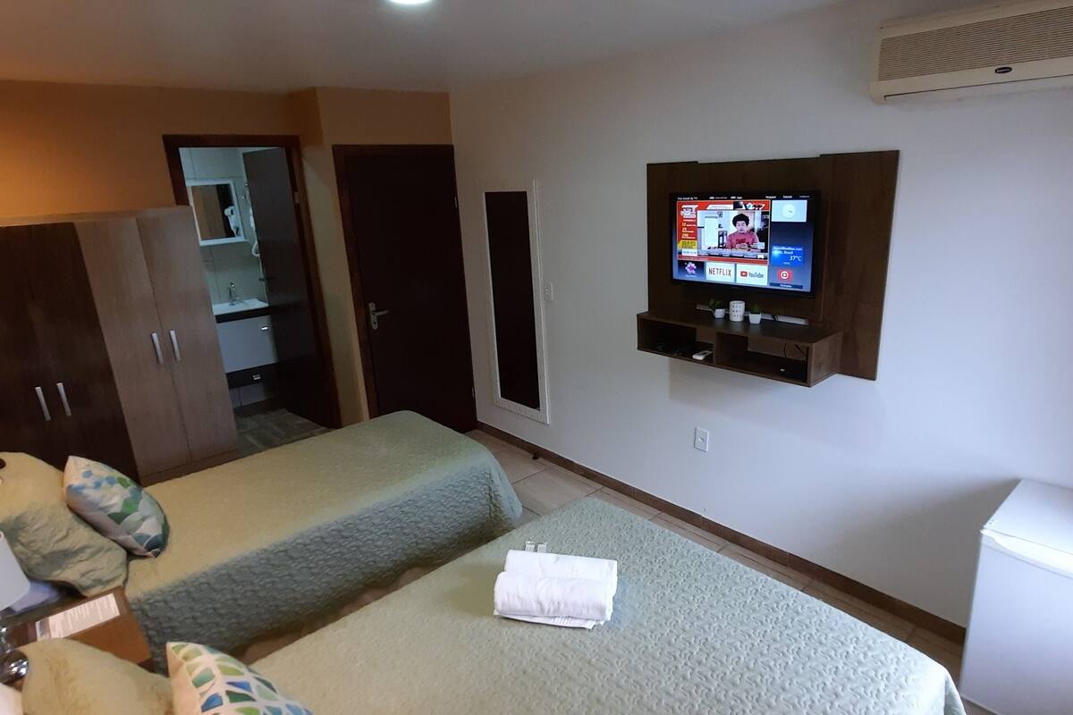 Une chambre d’hôtel avec deux lits, un téléviseur fixé au mur, une armoire, une petite table avec une lampe et un réfrigérateur.