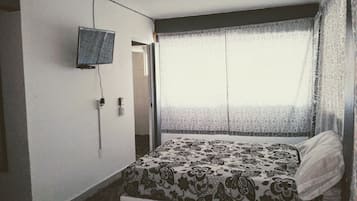 Habitación de hotel con cama, televisor montado en la pared y ventana con cortinas de gasa.