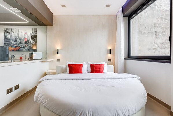 A modern hotel room with a large bed, two red pillows, a nightstand, a mirror, and a window with a view of the city.