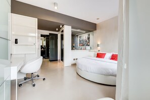 A modern hotel room with a white desk, a white chair, a curved bed with red pillows, and a cityscape mural on the wall.