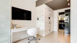 A modern, minimalist office space with a white desk, a white office chair, and a large flat-screen TV mounted on the wall.