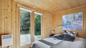 Camera da letto in una baita di legno, con un letto, un comodino, una lampada, un quadro e una finestra vista verde.