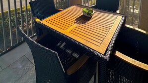 Un área de estar al aire libre con techo, una mesa de madera y sillas de mimbre negro, con vista a una calle con árboles y cielo despejado.