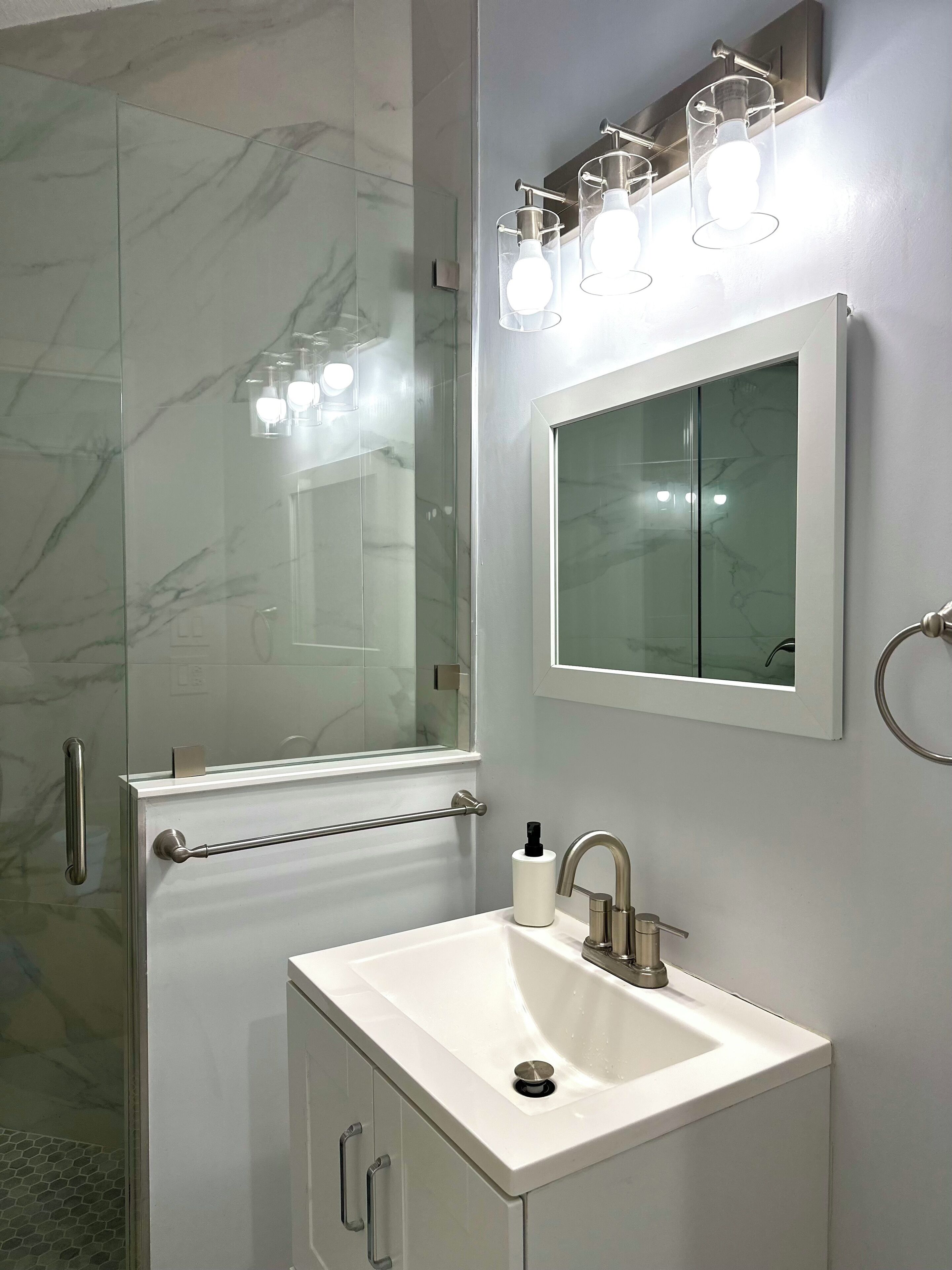 Un baño moderno con cabina de ducha de vidrio, un lavabo blanco con un solo grifo y un espejo con una lámpara encima.