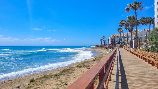 Un paseo marítimo de madera bordeando una playa, con palmeras y edificios al fondo.