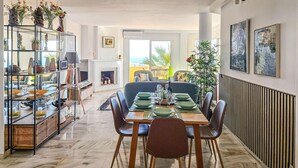 Un comedor con una mesa dispuesta para seis personas, rodeada de sillas. A la izquierda, una estantería con diversos objetos, y un amplio ventanal con vistas al exterior.