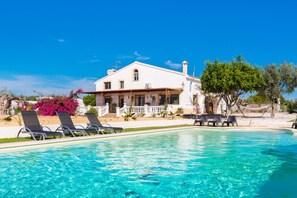 Una villa blanca con piscina, rodeada de vegetación y un cielo azul claro.