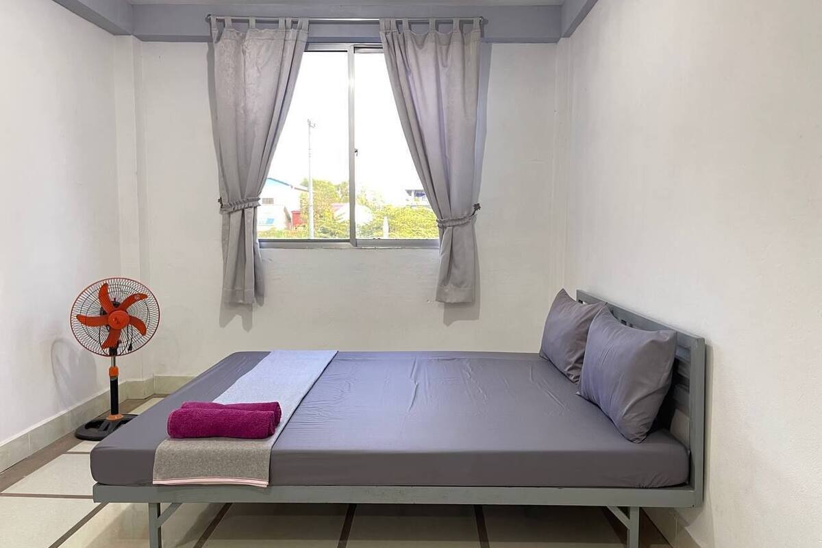 Une chambre avec un lit simple, un matelas gris, un ventilateur et une fenêtre avec des rideaux.