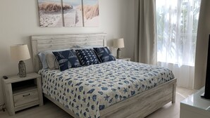 A bedroom with a wooden bed, two bedside tables with lamps, a large beach-themed painting above the bed, and a window with sheer curtains. - Luxury and charming 2Bdrm condo, minutes from beautiful Clearwater beach!  (Clearwater)