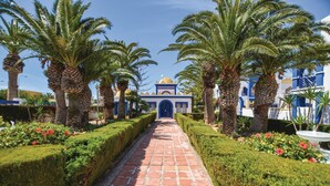 A pathway leading to a building with a blue facade and a golden dome, surrounded by palm trees and well-maintained hedges.