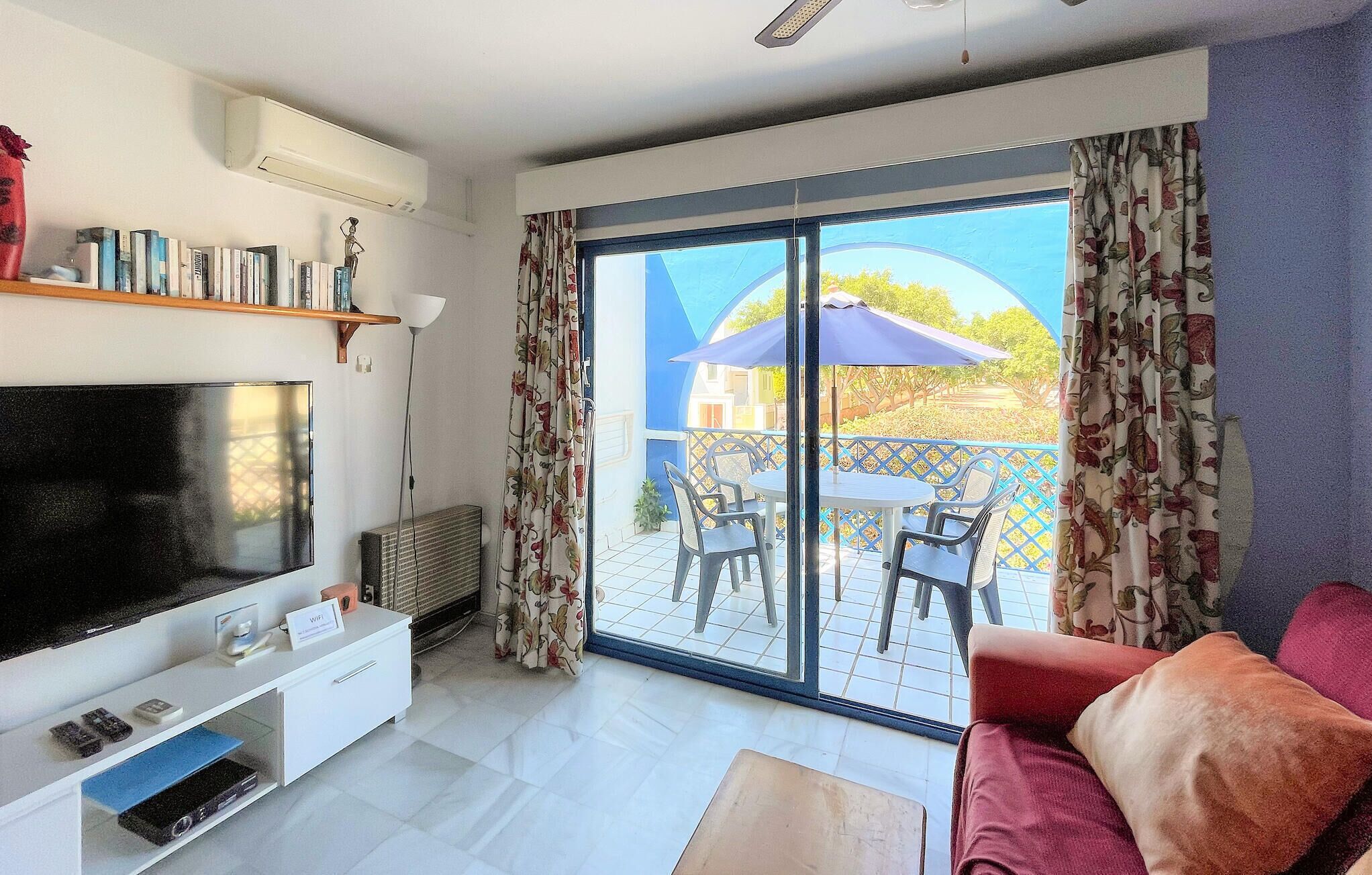A living room with a TV, white furniture, a balcony with chairs, and a view of the outside.