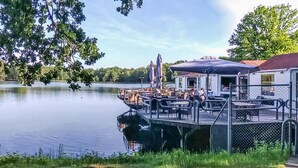 Un muelle junto al lago con mobiliario exterior y zona cubierta, rodeado de vegetación y árboles.