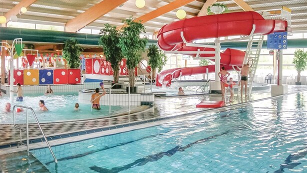 Piscina interior com várias pistas de natação, um escorrega aquático vermelho e área de recreação com estruturas coloridas.