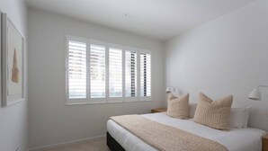 Un dormitorio con una cama grande, dos mesitas de noche, una ventana con persianas y una lámpara de pared.