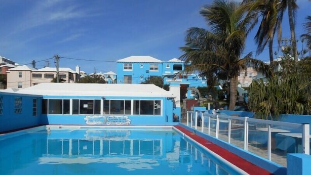 A swimming pool with a blue building adjacent to it, surrounded by palm trees and other buildings. - Blue Diamond Apartments (Warwick Parish)