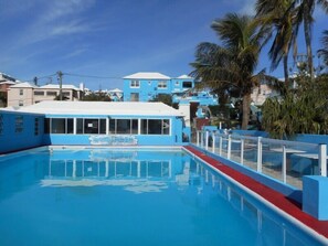 A swimming pool with a blue building adjacent to it, surrounded by palm trees and other buildings. - Blue Diamond Apartments (Warwick Parish)