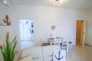 A living room with a sofa, a dining table with chairs, a kitchen in the background, and a door leading to another room. - Apartment summer wind - app. 1 - 1. Row on the beach and sea (Binz)