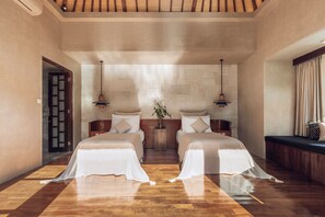 Habitación de hotel con dos camas, suelo de madera, pared con azulejos, banco de madera y lámparas colgantes.