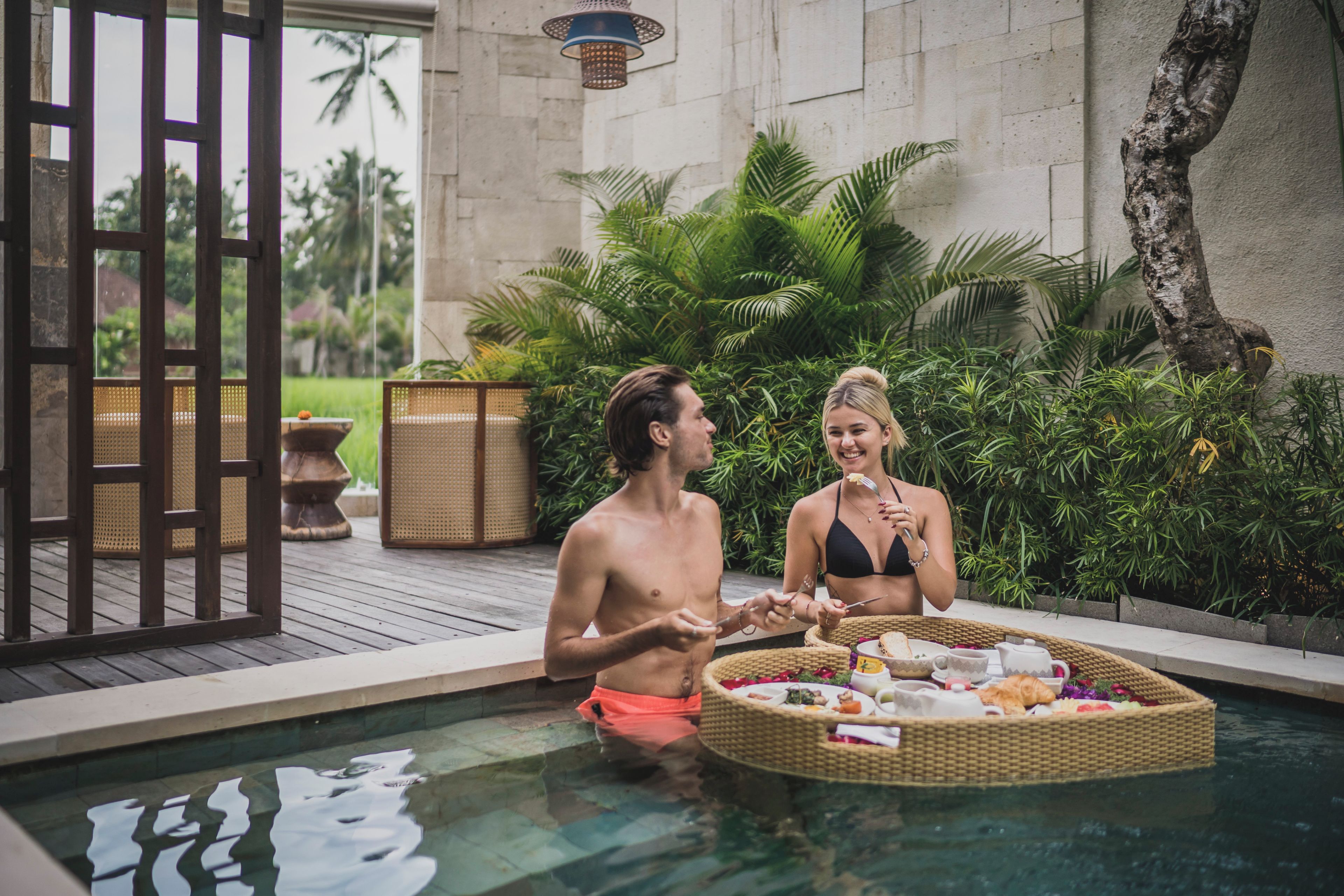 A poolside setting with a dining table and chairs, surrounded by lush greenery and a stone wall.