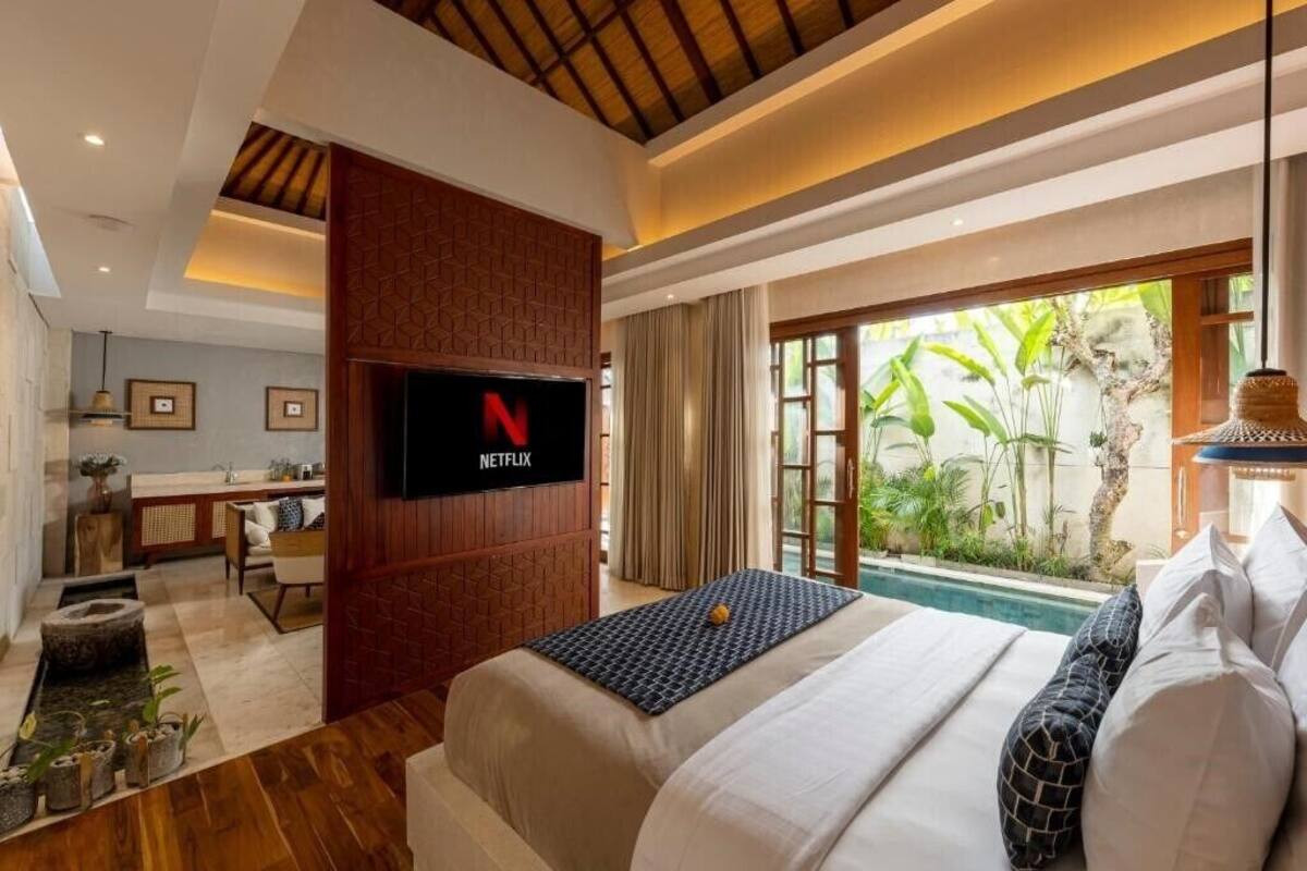 Ein modernes Hotelzimmer mit einem Bett, einem an einer Holzverkleidung montierten Fernseher, einem kleinen Schreibtisch und Blick auf Grün hinter großen Fenstern.