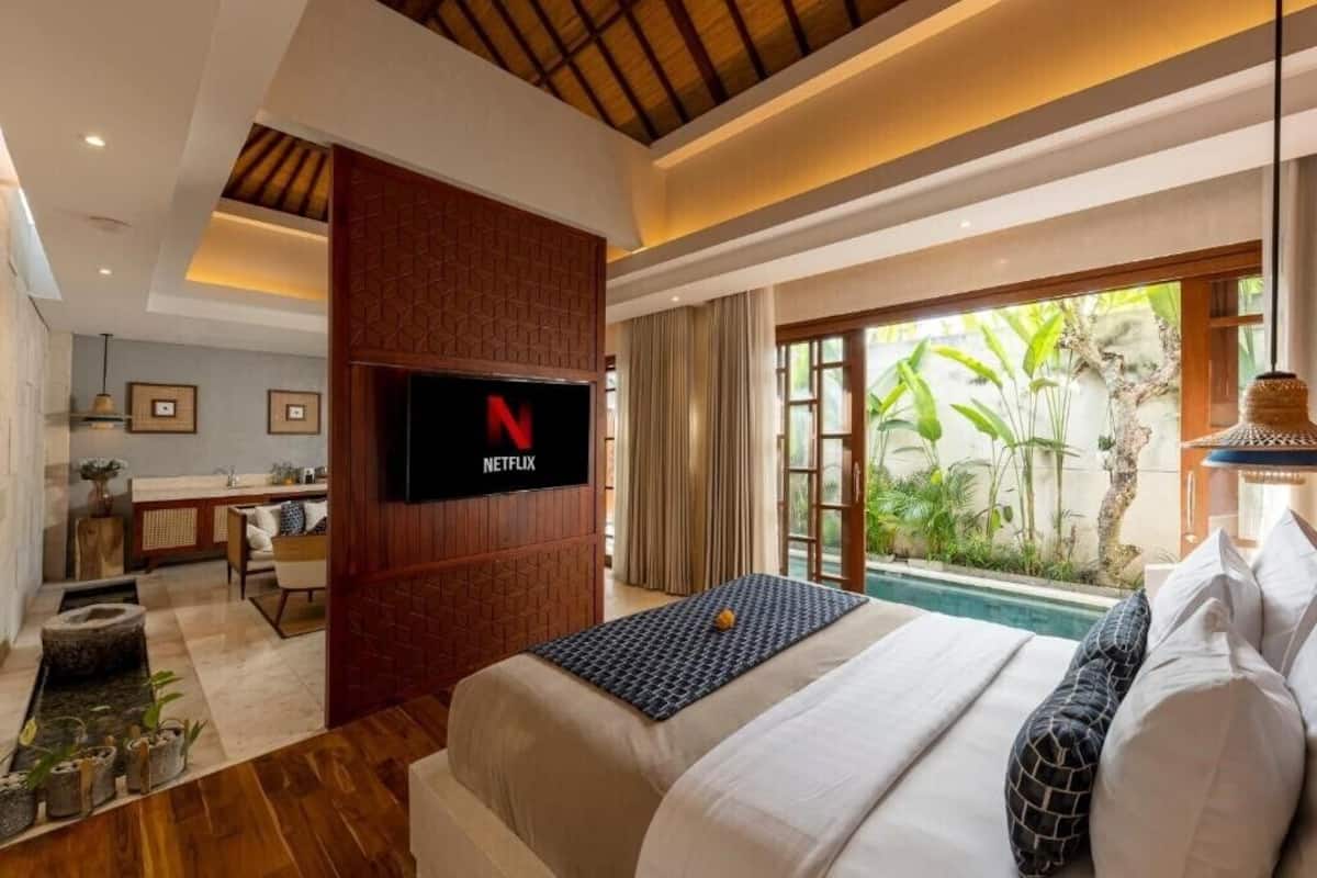 Una habitación de hotel moderna con una cama, un televisor montado en un panel de madera, un escritorio pequeño y vistas a zonas verdes a través de amplios ventanales.
