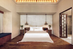 Un dormitorio amplio con una cama grande, dos mesitas de noche, un tocador, un banco de madera y una puerta que da al baño.