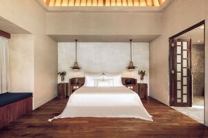 A spacious bedroom with a large bed, two bedside tables, a nightstand, a wooden bench, and a door leading to a bathroom.