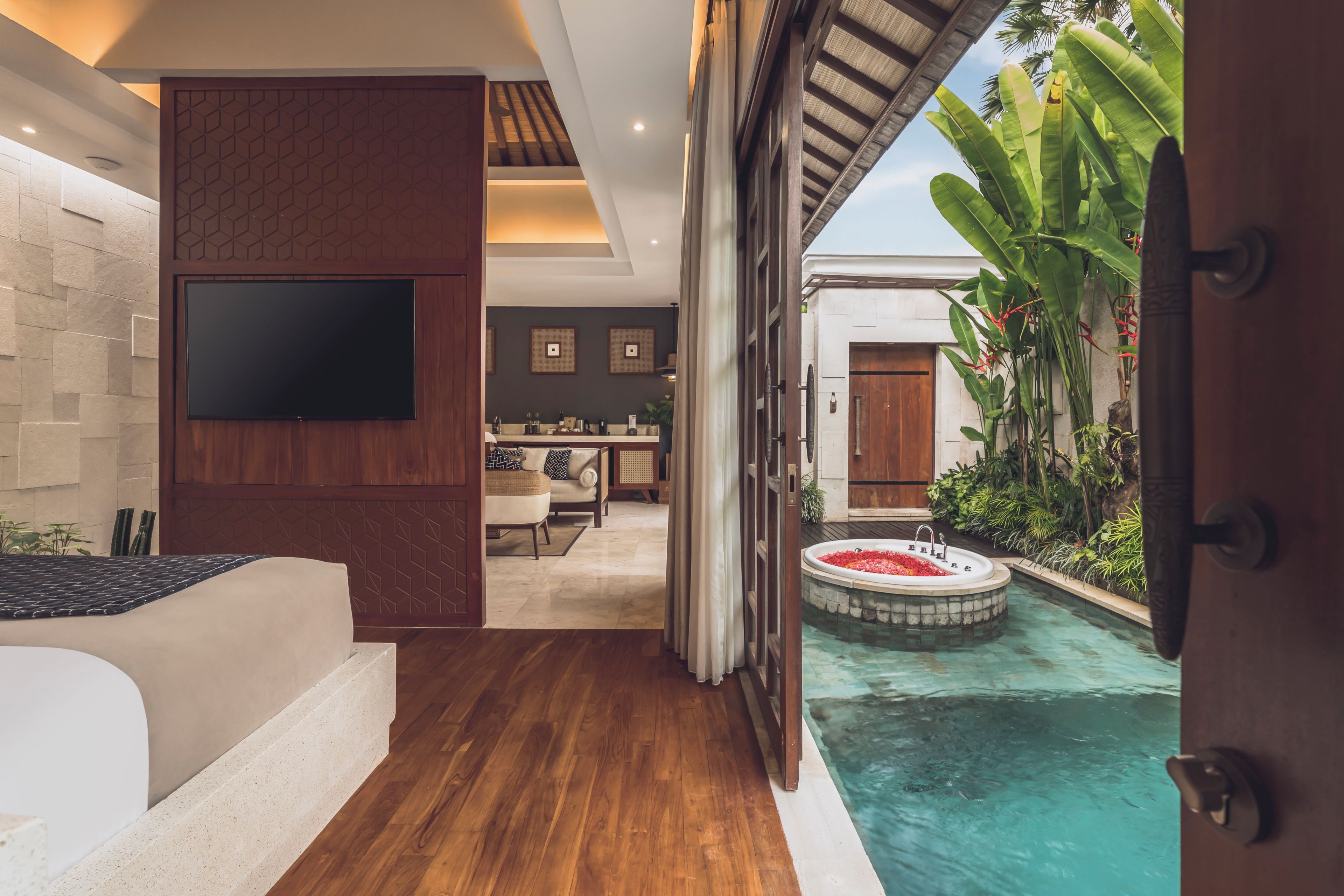 A modern interior with a flat-screen TV mounted on a wall, a wooden floor, a seating area with a sofa and a dining table, and a small pool with water lilies.