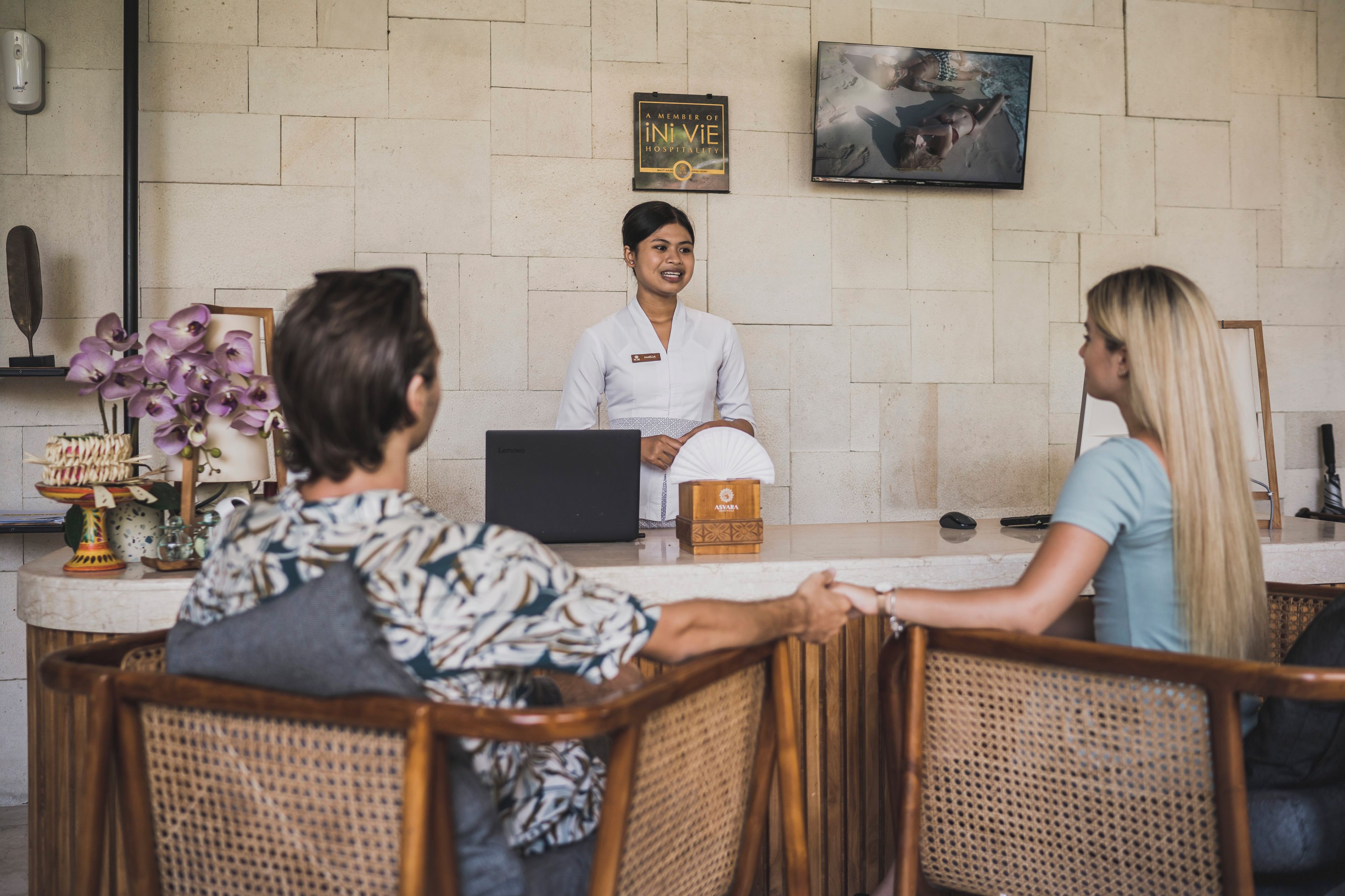 A reception desk with a laptop, a name tag, and a wooden box. There are wicker chairs and a wall-mounted TV.