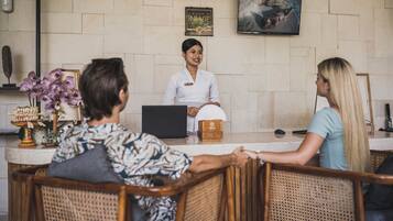 A reception desk with a laptop, a name tag, and a wooden box. There are wicker chairs and a wall-mounted TV.