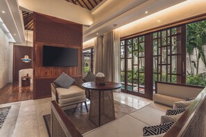 A modern living room with a flat-screen TV, comfortable seating, and large windows offering a view of greenery.