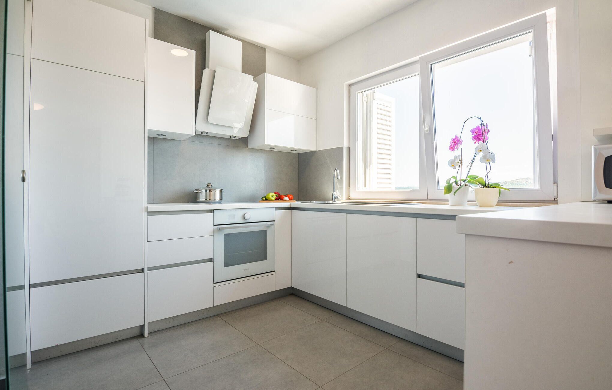 A modern kitchen with white cabinets, a built-in oven, and a window with potted orchids.