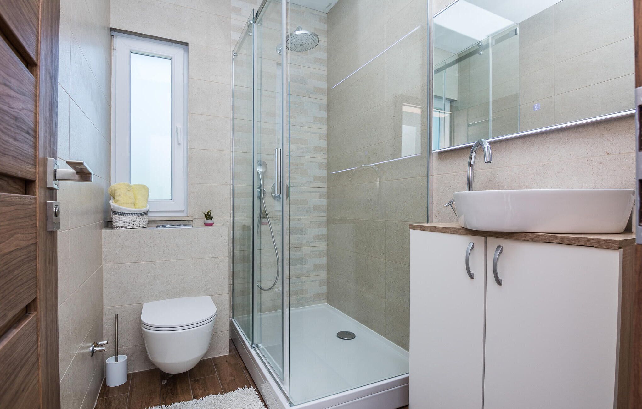 A modern bathroom with a glass shower enclosure, a white toilet, a sink with a wooden countertop, and a small window above the toilet.