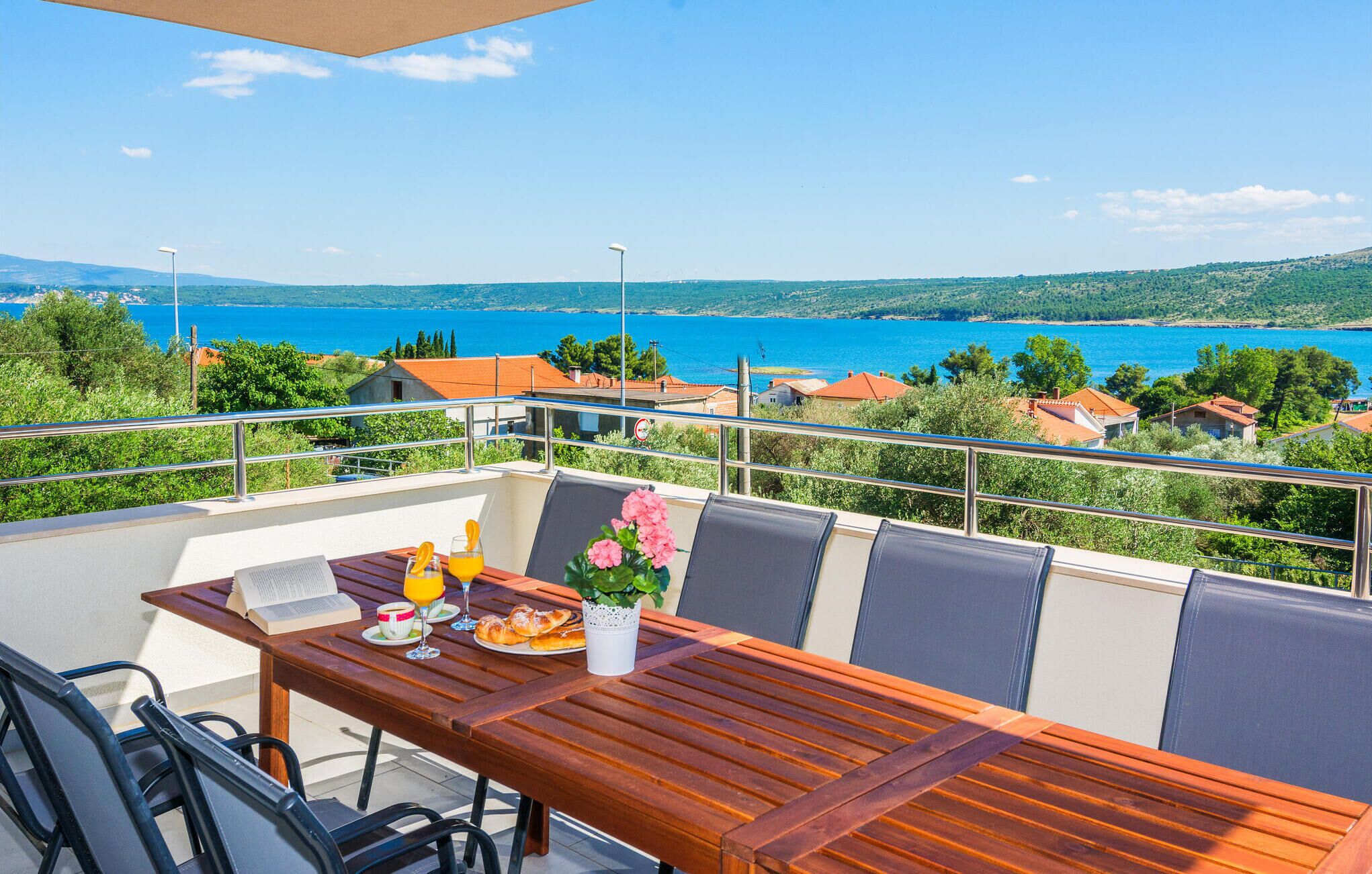 Varanda com mesa de madeira posta para o café da manhã, com vista para a paisagem costeira com casas e um mar azul e límpido.