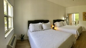 A hotel room with two beds, a desk, a chair, a potted plant, and a window with a view of greenery. - Sweet Spot Studio in PGA Village - 2 beds B (Port St Lucie)