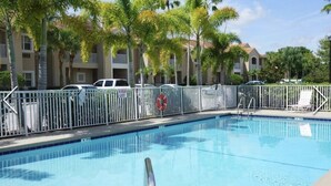A swimming pool with a metal fence, palm trees, and a building in the background. - Sweet Spot Studio in PGA Village - 2 beds B (Port St Lucie)