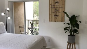 A bedroom with a bed, a potted plant, a wooden wall art, and a balcony with a chair and table. - Sweet Spot Studio in PGA Village - 2 beds B (Port St Lucie)