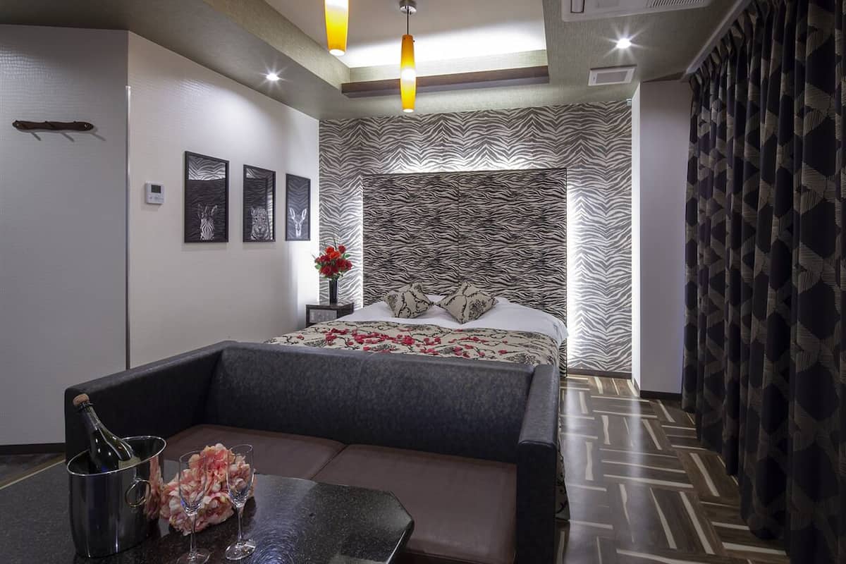 A modern hotel room with a bed, a sofa, a dining table with a bottle of champagne, and a vase of flowers.
