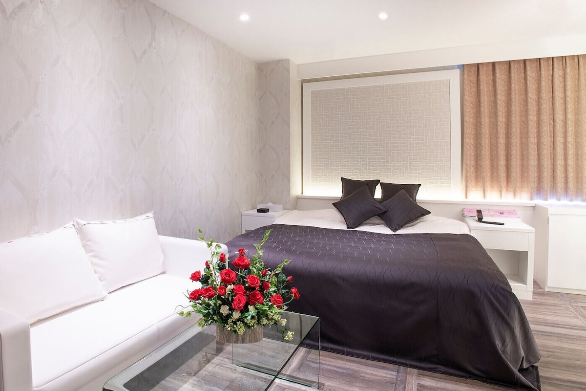 A modern hotel room with a white sofa, a dark bed, and a glass coffee table with a red flower arrangement.