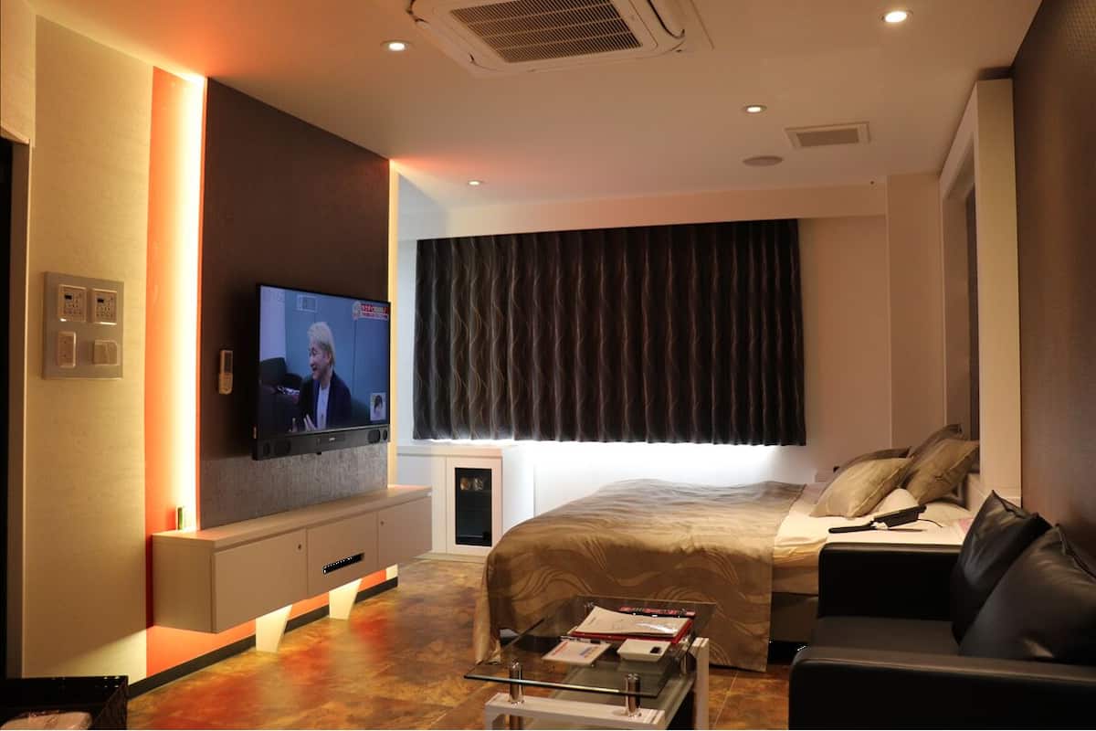 A modern hotel room with a large flat-screen TV mounted on the wall, a bed with a dark headboard, a sofa, and a small table with a tray.